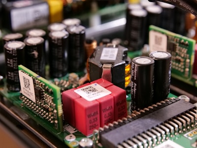 A close-up view of electronic components on a circuit board, featuring capacitors, resistors, and microchips. The arrangement includes cylindrical capacitors alongside rectangular chips with QR codes and other components connected by intricate wiring.