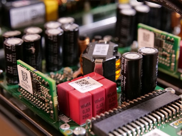 A close-up view of electronic components on a circuit board, featuring capacitors, resistors, and microchips. The arrangement includes cylindrical capacitors alongside rectangular chips with QR codes and other components connected by intricate wiring.