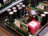 A close-up view of electronic components on a circuit board, including capacitors, inductors, and semiconductor chips. The components are densely packed, with visible QR codes on some of the parts. The circuit board is part of a larger electronic device with a metallic enclosure.