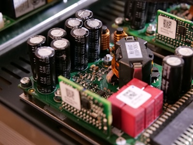 A close-up view of electronic components on a circuit board, including capacitors, inductors, and semiconductor chips. The components are densely packed, with visible QR codes on some of the parts. The circuit board is part of a larger electronic device with a metallic enclosure.