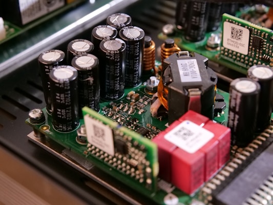 A close-up view of electronic components on a circuit board, including capacitors, inductors, and semiconductor chips. The components are densely packed, with visible QR codes on some of the parts. The circuit board is part of a larger electronic device with a metallic enclosure.
