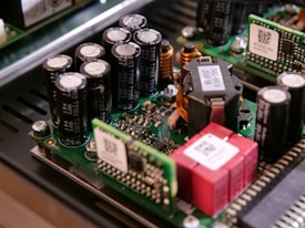 A close-up view of electronic components on a circuit board, including capacitors, inductors, and semiconductor chips. The components are densely packed, with visible QR codes on some of the parts. The circuit board is part of a larger electronic device with a metallic enclosure.