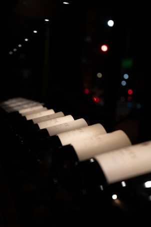 A row of wine bottles displayed in a dimly lit environment, with a focus on the soft lighting illuminating the labels. The background features blurred city lights, adding a touch of vibrancy.