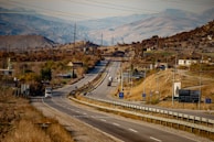 A winding road stretches into the distance through a hilly landscape with scattered houses and utility poles. Several vehicles are visible on the road, including trucks and cars. The terrain is primarily dry and brown, with patches of greenery and rocky hills in the background. Road signs and billboards are present along the roadside.
