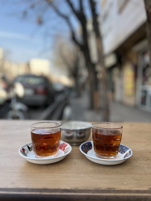 Two glass cups of tea are placed on a wooden table, each sitting on a decorative saucer. The background is blurred, showing an outdoor setting with trees and a street. The atmosphere suggests a casual outdoor setting.