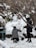 Two people are sculpting a snow figure in a snowy outdoor setting surrounded by bare trees. One person is kneeling and shaping the snow, while the other stands nearby observing. The area is covered in snow, with some foliage visible.