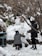 Two people are sculpting a snow figure in a snowy outdoor setting surrounded by bare trees. One person is kneeling and shaping the snow, while the other stands nearby observing. The area is covered in snow, with some foliage visible.