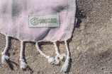 A woven fabric with a pink hue and white fringe is partially spread across a sandy surface. A label with 'Sand Cloud' is sewn onto the fabric, accompanied by a circular logo.