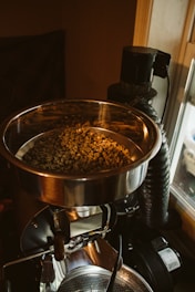 A metal container filled with unroasted coffee beans is part of a coffee roasting machine in a dimly lit setting. The focus is on the beans and the reflective surface of the container.