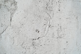 A textured cement wall with rough surfaces and subtle cracks. The surface has visible patterns and imperfections that create an uneven appearance.