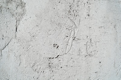 A textured cement wall with rough surfaces and subtle cracks. The surface has visible patterns and imperfections that create an uneven appearance.