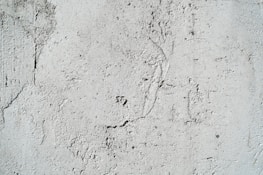 A textured cement wall with rough surfaces and subtle cracks. The surface has visible patterns and imperfections that create an uneven appearance.