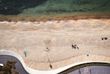 Aerial view of a sandy beach bordered by clear greenish water and a curving boardwalk. Groups of people are scattered across the sand, some sitting close together on colorful blankets. The beach shows footprints and smooth sand. Nearby are patches of greenery and a pathway with shadows from overhead lights and a tree.