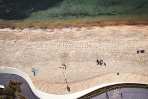 Aerial view of a sandy beach bordered by clear greenish water and a curving boardwalk. Groups of people are scattered across the sand, some sitting close together on colorful blankets. The beach shows footprints and smooth sand. Nearby are patches of greenery and a pathway with shadows from overhead lights and a tree.