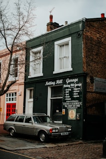 A vintage car is parked in front of a small, dark green building with a sign that reads 'Notting Hill Garage.' The building features white-trimmed windows and an array of repair services listed on the front. A leafless tree stands nearby, and the surrounding architecture has a classic, urban appearance.