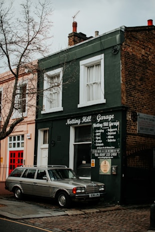A vintage car is parked in front of a small, dark green building with a sign that reads 'Notting Hill Garage.' The building features white-trimmed windows and an array of repair services listed on the front. A leafless tree stands nearby, and the surrounding architecture has a classic, urban appearance.