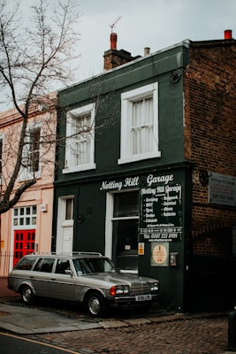 A vintage car is parked in front of a small, dark green building with a sign that reads 'Notting Hill Garage.' The building features white-trimmed windows and an array of repair services listed on the front. A leafless tree stands nearby, and the surrounding architecture has a classic, urban appearance.