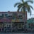 A colorful mural featuring a woman's face with vibrant makeup and headwrap decorates the upper part of a building. The mural is accompanied by images of fruits like oranges and lemons, as well as tropical plants and a rooster. Below the mural, a sign reads 'Party Cake Bakery' with a line of people standing on the sidewalk in front. A tall palm tree stands to the right of the building.