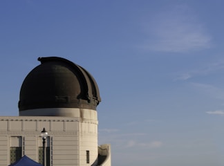 A large observatory dome with its dark top contrasting against a clear blue sky. The building features architectural details like decorative patterns and a small lamp post is visible in the foreground.