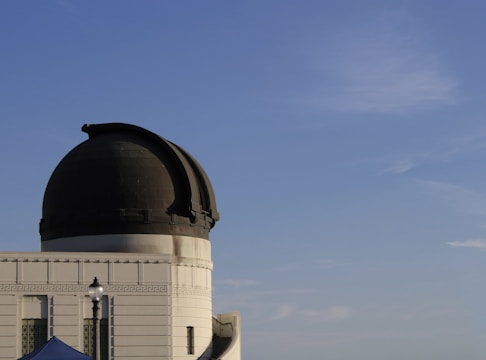 A large observatory dome with its dark top contrasting against a clear blue sky. The building features architectural details like decorative patterns and a small lamp post is visible in the foreground.