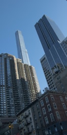 A cityscape featuring several tall skyscrapers against a clear blue sky. The buildings vary in architectural style, with modern glass structures juxtaposed with older brick architecture. The scene conveys a sense of urban density and architectural diversity.