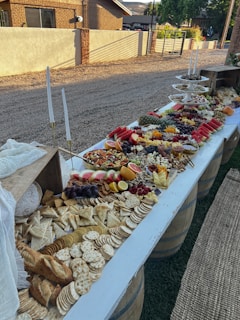 A long, elegantly arranged table features an abundant assortment of food, including crackers, cheeses, breads, fruits like watermelon and oranges, and various nuts and pretzels. This outdoor setup is placed over wine barrels with white tableware and candles adding to its festive charm.