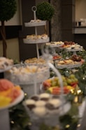 A lavish buffet setup featuring tiered trays filled with various sweets and appetizers. The trays contain cupcakes, fruits like grapes and watermelon, and small desserts. Decorative greenery and lights adorn the table, adding a festive atmosphere. Two topiary trees are visible in the background, enhancing the elegant presentation.