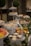 A lavish buffet setup featuring tiered trays filled with various sweets and appetizers. The trays contain cupcakes, fruits like grapes and watermelon, and small desserts. Decorative greenery and lights adorn the table, adding a festive atmosphere. Two topiary trees are visible in the background, enhancing the elegant presentation.