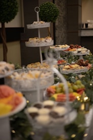 A lavish buffet setup featuring tiered trays filled with various sweets and appetizers. The trays contain cupcakes, fruits like grapes and watermelon, and small desserts. Decorative greenery and lights adorn the table, adding a festive atmosphere. Two topiary trees are visible in the background, enhancing the elegant presentation.