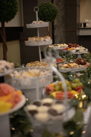 A lavish buffet setup featuring tiered trays filled with various sweets and appetizers. The trays contain cupcakes, fruits like grapes and watermelon, and small desserts. Decorative greenery and lights adorn the table, adding a festive atmosphere. Two topiary trees are visible in the background, enhancing the elegant presentation.