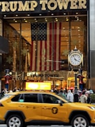 The entrance to a large building is adorned with a massive American flag and features gold-toned lettering above the entrance. A traditional clock is situated on the right, with decorative figures of soldiers on either side. A yellow taxi is driving past, while a crowd of people gathers near the entrance.