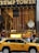 The entrance to a large building is adorned with a massive American flag and features gold-toned lettering above the entrance. A traditional clock is situated on the right, with decorative figures of soldiers on either side. A yellow taxi is driving past, while a crowd of people gathers near the entrance.