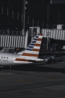 An aircraft with a distinctive striped tail parked at an airport gate, equipped with a jet bridge. The surrounding area includes various ground service vehicles and equipment. The environment is dimly lit, suggesting that it might be early morning or late evening.