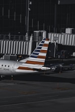 An aircraft with a distinctive striped tail parked at an airport gate, equipped with a jet bridge. The surrounding area includes various ground service vehicles and equipment. The environment is dimly lit, suggesting that it might be early morning or late evening.
