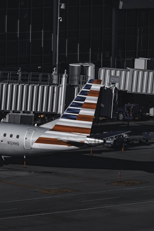 An aircraft with a distinctive striped tail parked at an airport gate, equipped with a jet bridge. The surrounding area includes various ground service vehicles and equipment. The environment is dimly lit, suggesting that it might be early morning or late evening.