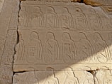 An ancient stone wall features detailed carved images and hieroglyphics, displaying figures in profile wearing traditional Egyptian headdresses. The carvings are arranged in a repetitive pattern, reflecting the artistry of ancient Egyptian culture.