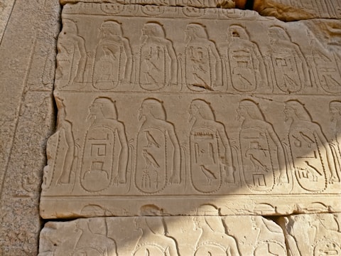 An ancient stone wall features detailed carved images and hieroglyphics, displaying figures in profile wearing traditional Egyptian headdresses. The carvings are arranged in a repetitive pattern, reflecting the artistry of ancient Egyptian culture.