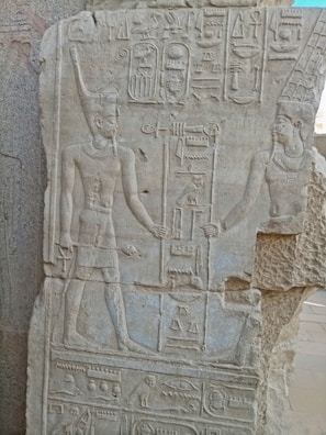 An ancient stone carving featuring two figures adorned with traditional Egyptian attire and headdresses, with intricate hieroglyphs surrounding them. The figures appear to hold ceremonial staffs, and the entire scene is carved into a light-colored stone surface.