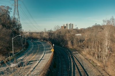 A curving road with a few cars runs parallel to train tracks, set against a backdrop of trees and utility poles. In the distance, tall buildings rise above the landscape. The area appears to be an urban edge, transitioning from a wooded setting to a city environment.