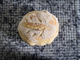 A round loaf of homemade bread rests on a cooling rack. The bread has a golden-brown crust dusted with flour and textured surface showing artisanal slashes.