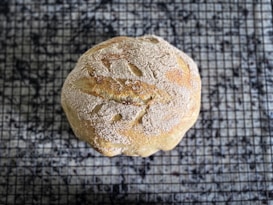 A round loaf of homemade bread rests on a cooling rack. The bread has a golden-brown crust dusted with flour and textured surface showing artisanal slashes.