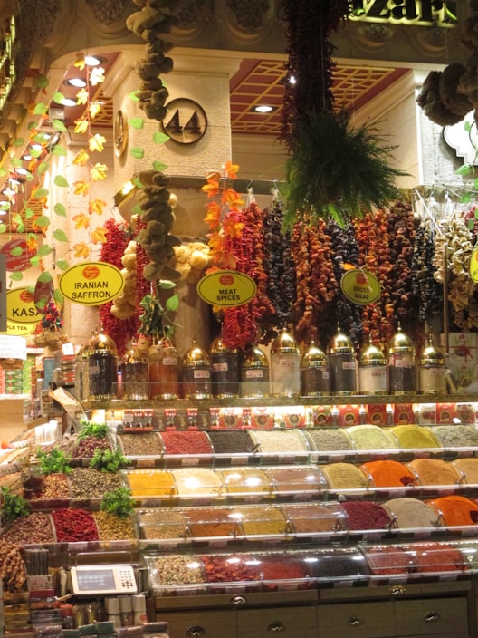 A vibrant spice market display featuring an array of colorful spices in clear containers. The backdrop includes hanging decorations made of dried herbs, garlic, and peppers. Labels such as 'Iranian Saffron' and 'Meat Spices' are visible. The ambiance is warm and inviting, with intricate architectural details and decorative lighting.