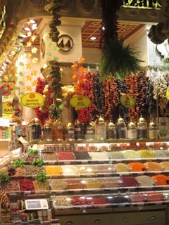 A vibrant spice market display featuring an array of colorful spices in clear containers. The backdrop includes hanging decorations made of dried herbs, garlic, and peppers. Labels such as 'Iranian Saffron' and 'Meat Spices' are visible. The ambiance is warm and inviting, with intricate architectural details and decorative lighting.
