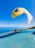 A bright yellow and blue paraglider takes off from a launch pad overlooking a vast expanse of blue ocean. Several people assist in preparing for the flight, with a clear blue sky above them and distant islands on the horizon.