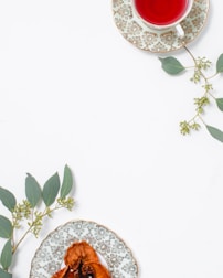 A cup of red herbal tea sits on an ornate plate with a floral pattern, placed alongside green eucalyptus leaves. A second decorative plate holds dried fruit, also adorned with eucalyptus sprigs, arranged on a clean, white surface.