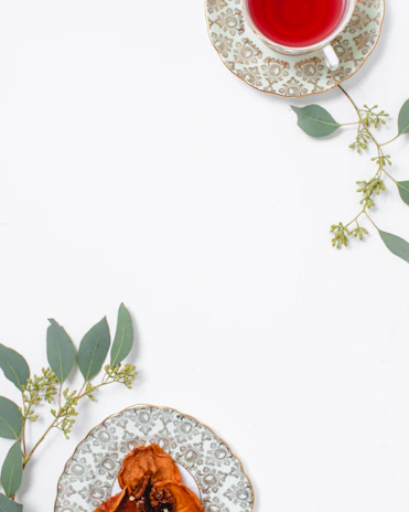 A cup of red herbal tea sits on an ornate plate with a floral pattern, placed alongside green eucalyptus leaves. A second decorative plate holds dried fruit, also adorned with eucalyptus sprigs, arranged on a clean, white surface.
