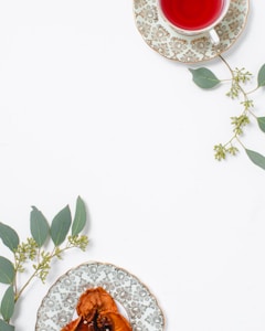 A cup of red herbal tea sits on an ornate plate with a floral pattern, placed alongside green eucalyptus leaves. A second decorative plate holds dried fruit, also adorned with eucalyptus sprigs, arranged on a clean, white surface.