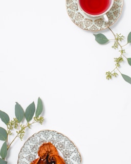 A cup of red herbal tea sits on an ornate plate with a floral pattern, placed alongside green eucalyptus leaves. A second decorative plate holds dried fruit, also adorned with eucalyptus sprigs, arranged on a clean, white surface.