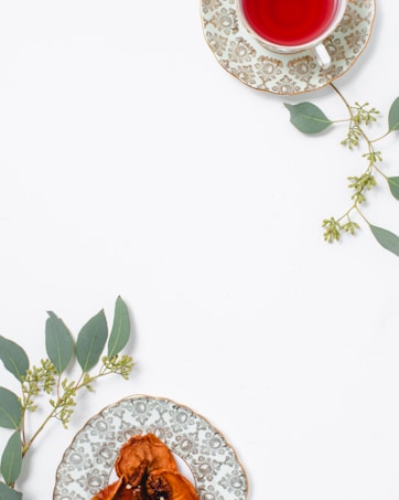 A cup of red herbal tea sits on an ornate plate with a floral pattern, placed alongside green eucalyptus leaves. A second decorative plate holds dried fruit, also adorned with eucalyptus sprigs, arranged on a clean, white surface.