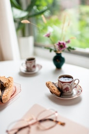 Elegant tea cups filled with dark coffee are placed on saucers on a white surface. A piece of biscotti rests beside one of the cups. In the background, a small green vase holds purple flowers, with greenery visible through a nearby window. Nearby, a pair of eyeglasses rests on a book or notebook.